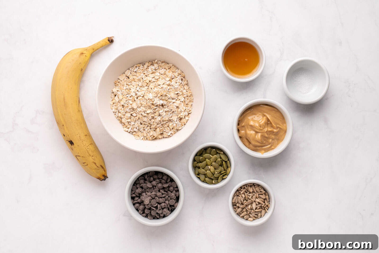 A banana, a bowl of rolled oats, and small bowls of honey, peanut butter, salt, pumpkin seeds, chocolate chips, and sunflower seeds are arranged on a white marble surface.