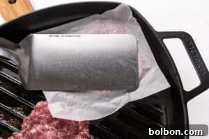 A metal spatula presses ground meat on parchment paper in a cast iron grill pan, partially flattening the meat for cooking.