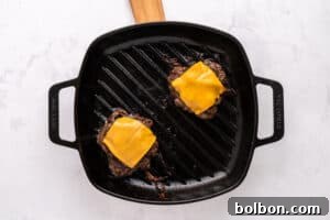 A black grill pan with two cooked hamburger patties, each topped with a slice of melted cheddar cheese, sits on a light-colored surface. The handle of the grill pan is wooden.