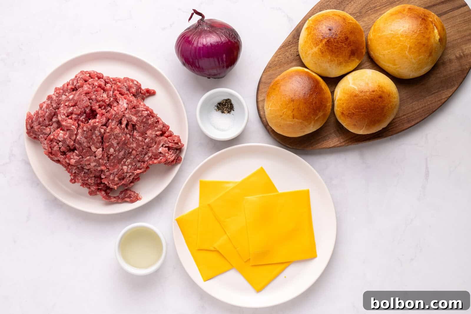 Top-down view of burger ingredients: a plate of ground beef, a red onion, salt and pepper, four burger buns on a wooden board, American cheese slices on a plate, and a small bowl of oil on a white surface.