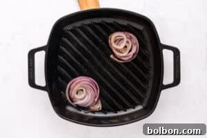 A black cast iron grill pan with two slices of red onion being grilled, one near the top right and one near the bottom left, on a white surface.