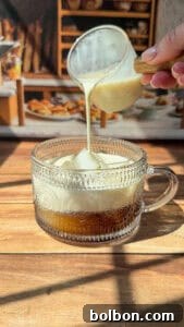 A hand pours creamy milk from a small glass pitcher into a patterned glass mug filled with coffee, set on a wooden surface with sunlight streaming in.