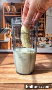 A hand holding half a peeled banana is about to place it into a blender cup partially filled with milk, on a wooden surface with a kitchen in the background.