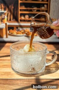 A hand pours espresso from a small glass pitcher over a scoop of vanilla ice cream in a decorative glass cup, creating an affogato. The scene is sunlit with baked goods and shelves in the blurred background.