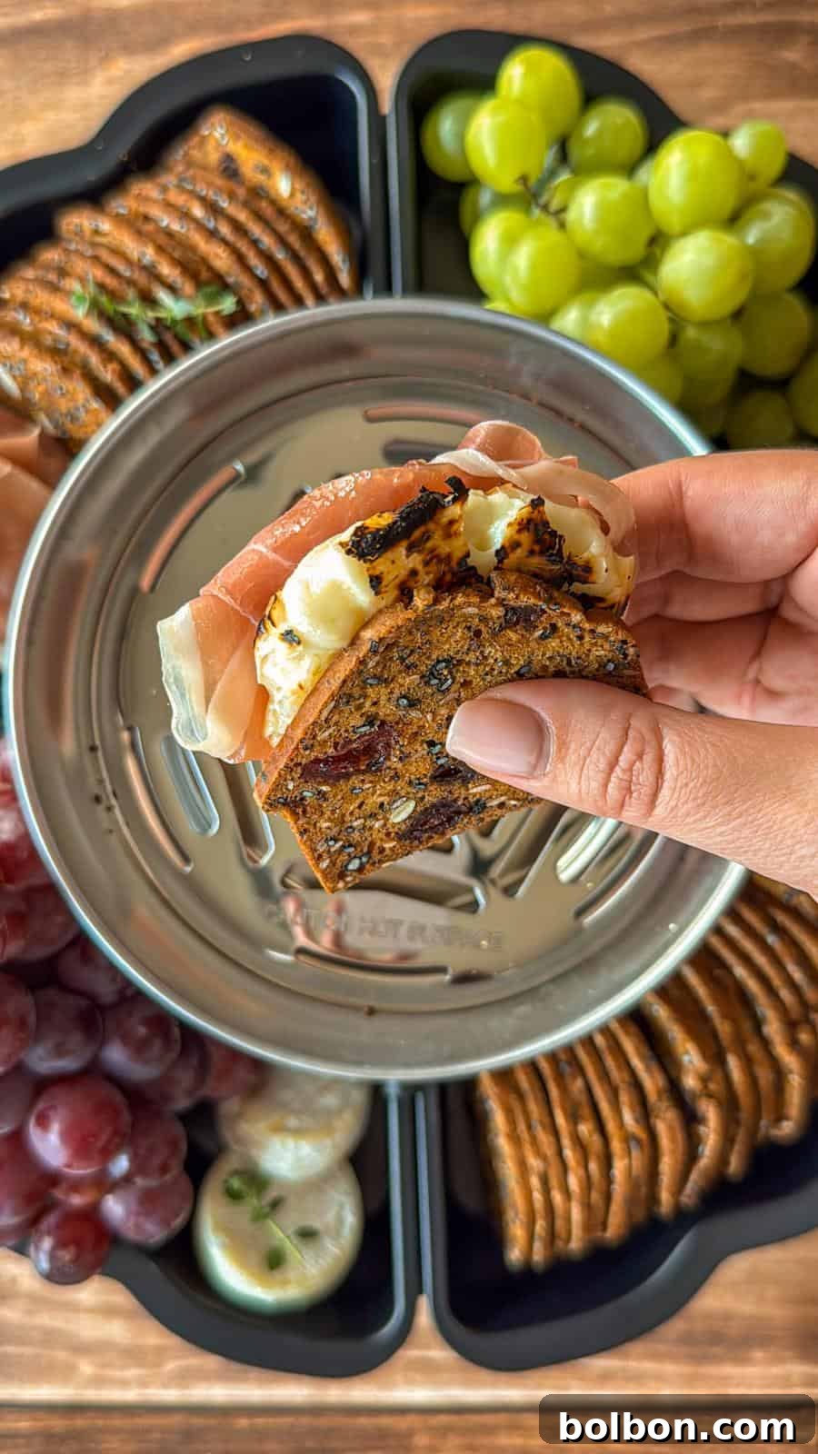 A hand holds a stack of crackers with cheese and prosciutto above a metal dish. The background shows green grapes, red grapes, crackers, and cheese displayed on a wooden surface.
