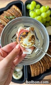 A hand holds a cracker topped with prosciutto and torched goat cheese over a serving tray with grapes, crackers, and cheese.