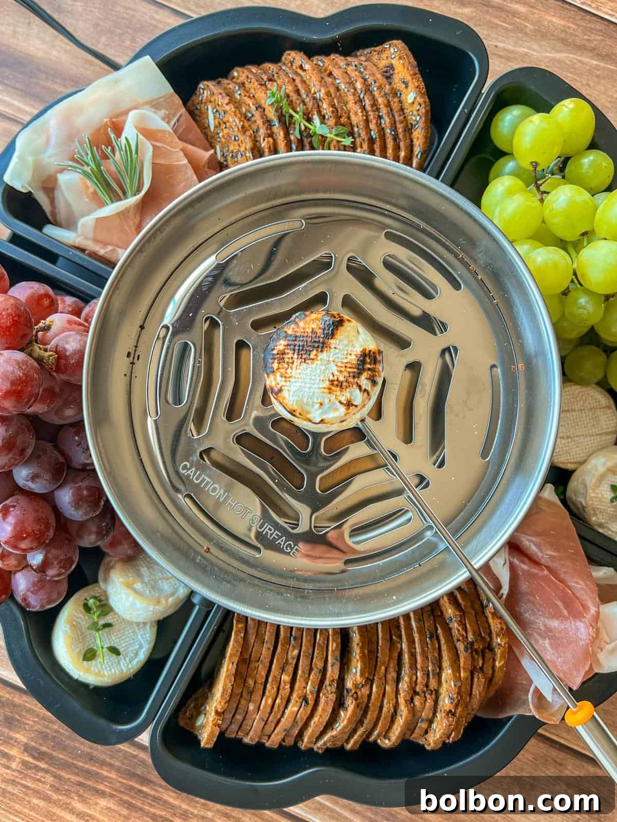 A metal burner toasts a marshmallow for Savory Smores on a skewer, surrounded by crackers, grapes, cheese, prosciutto, and herbs, all arranged in a black snack tray on a wooden surface.