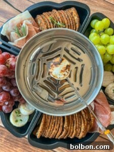 A metal burner toasts a marshmallow on a skewer, surrounded by crackers, grapes, banana slices, prosciutto, and herbs, all arranged in a black snack tray on a wooden surface.