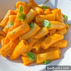 A white plate of rigatoni pasta coated in creamy orange Hidden Veggie Pasta Sauce, garnished with fresh basil leaves, with a gold fork resting on the side.