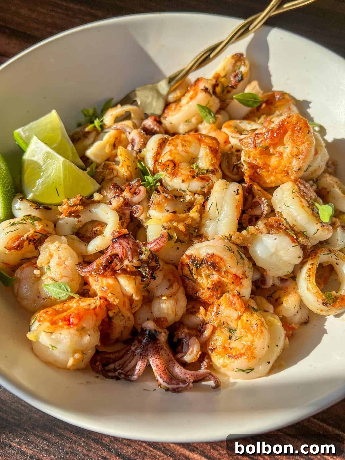 A white bowl filled with grilled shrimp and calamari, garnished with herbs and served with lime wedges, sits on a wooden surface in bright natural light.