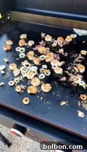 Seafood including shrimp and calamari rings cooking on a flat-top Blackstone grill with scattered seasoning and oil visible on the hot surface.