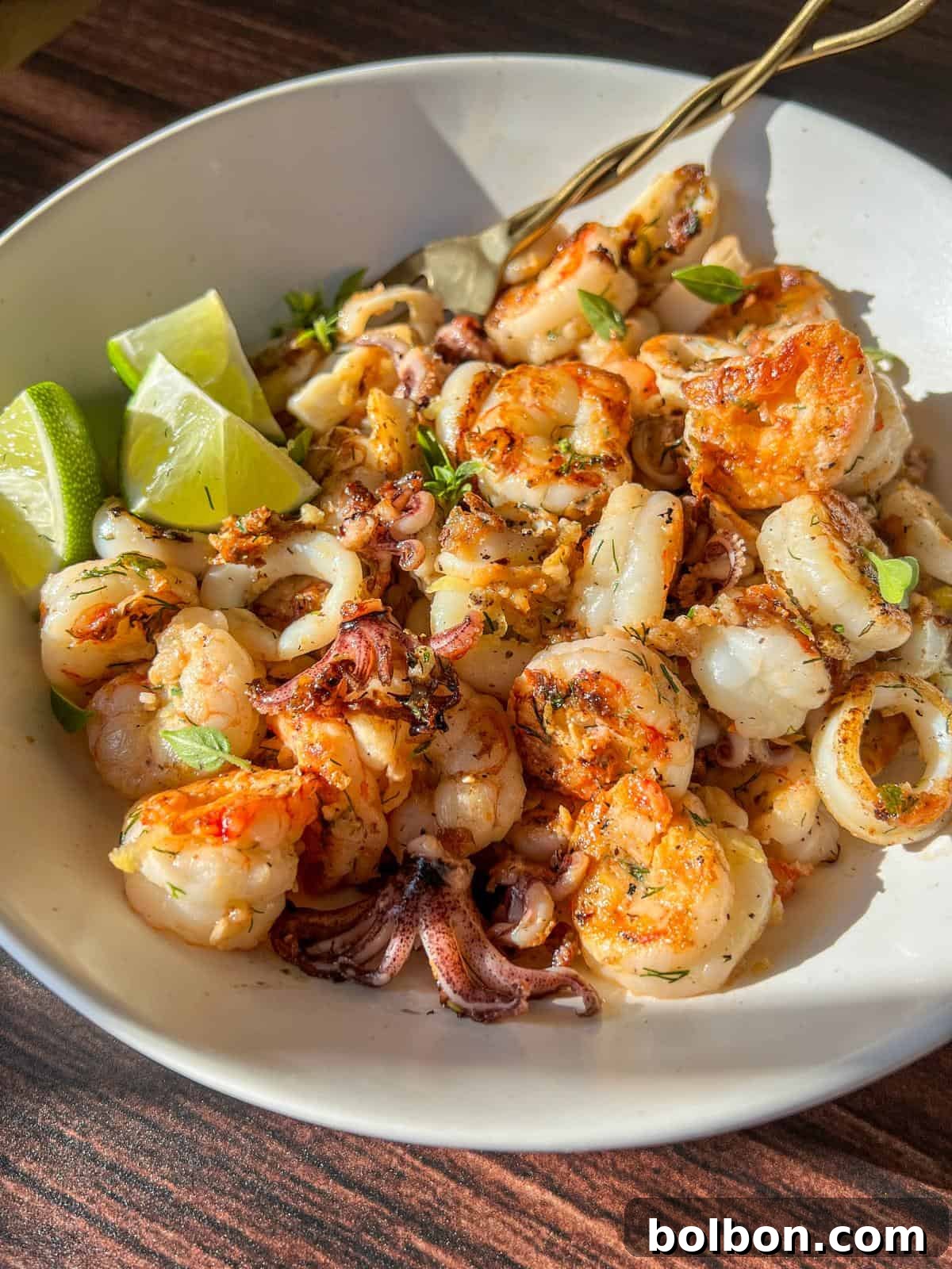 A bowl of grilled seafood, including shrimp and squid, garnished with fresh herbs and served with lime wedges, sits on a wooden table in bright sunlight.