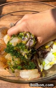 A hand mixes raw shrimp with chopped herbs, lemon zest, and seasonings in a glass bowl on a wooden surface.