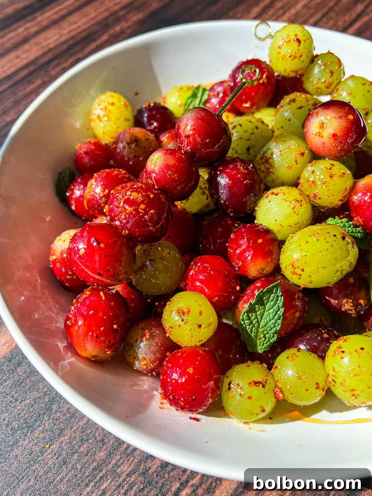 Chilled Tangy Grape Bites 2 A pristine white bowl overflowing with vibrant red and green grapes, generously coated in a shimmering layer of Tajin powder and aromatic seasoning. The bowl is elegantly garnished with fresh mint leaves and set against a rustic wooden backdrop, inviting you to discover its zesty allure.