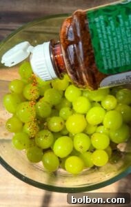 A bottle of chili powder is being sprinkled over a glass bowl filled with fresh green grapes on a wooden surface.
