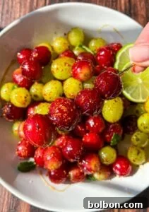A close-up of a bowl filled with glossy red and green gooseberries coated in spices, with a lime slice visible. A hand holds a skewer with three spiced gooseberries above the bowl.