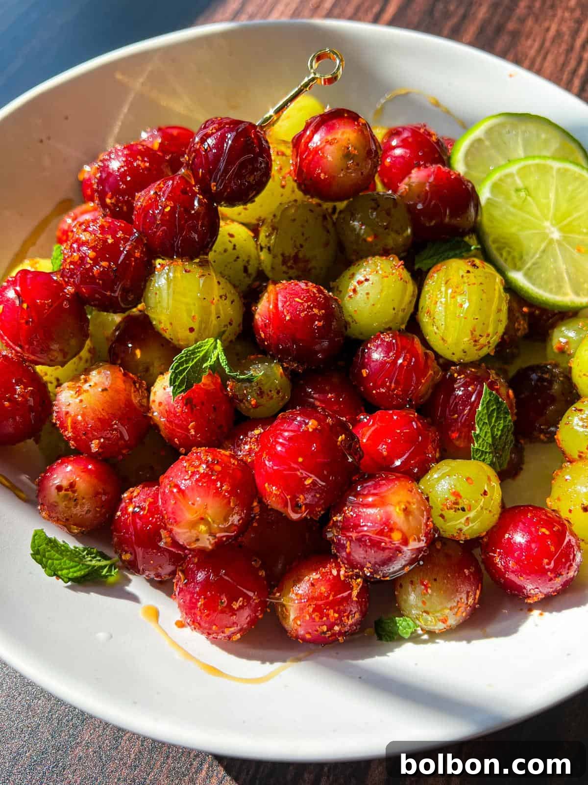 Chilled Tangy Grape Bites 3 A pristine white bowl filled with glossy red and green gooseberries, generously sprinkled with aromatic spices. The bowl is beautifully garnished with vibrant fresh mint leaves and delicate lime slices, resting elegantly on a rich, dark wooden surface.