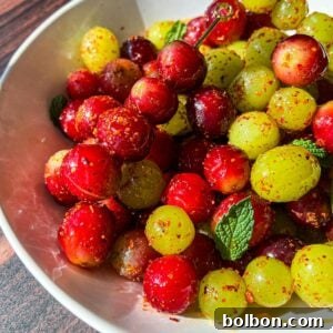 A white bowl filled with vibrant red and green grapes, generously coated in chili powder and seasoning, elegantly garnished with fresh mint leaves, all set on a rustic wooden surface.