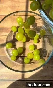 Green grapes are being poured into a clear glass bowl on a wooden surface, with sunlight casting shadows across the scene.