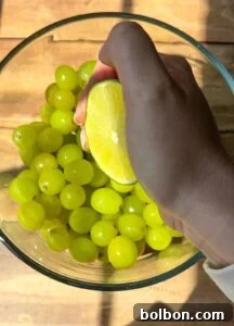 A hand squeezes a halved lime over a glass bowl filled with green grapes on a wooden surface. Sunlight casts shadows across the scene.