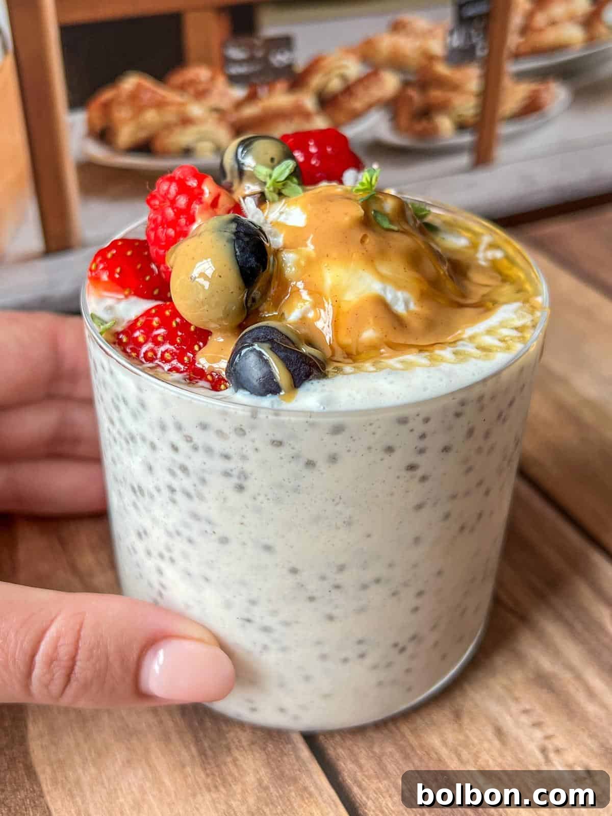 A glass of chia pudding topped with fresh strawberries, blueberries, a golden berry, and a generous dollop of peanut butter. A hand is holding the glass on a wooden table, with pastries blurred in the background.