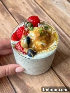 A hand holds a glass of chia pudding topped with strawberries, blueberries, gooseberries, a dollop of peanut butter, honey, and a sprig of fresh herbs, set on a wooden surface.