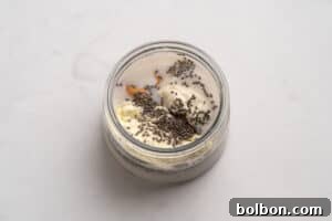 A glass jar filled with milk, chia seeds, a scoop of protein powder, and a dollop of peanut butter on a light, smooth surface.