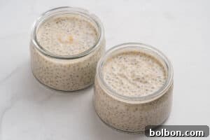 Two small glass jars filled with creamy chia seed pudding are placed on a white marble surface. The pudding is light beige with visible chia seeds evenly dispersed throughout.