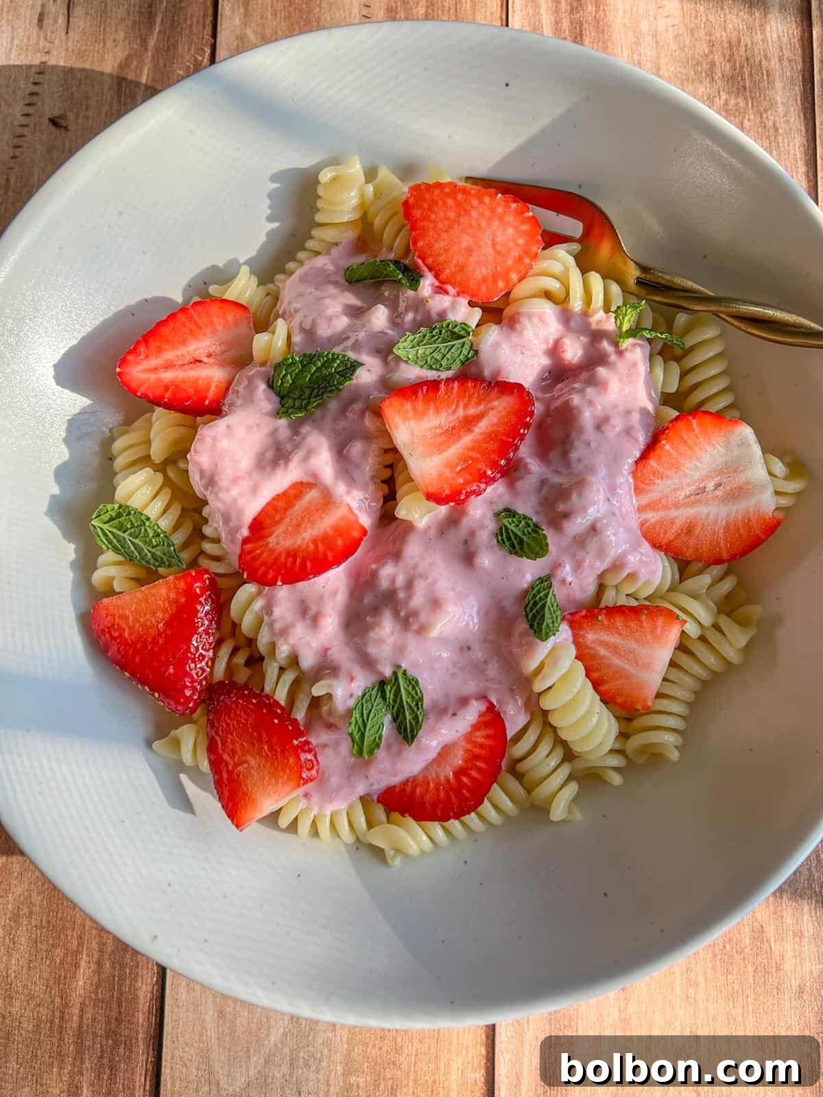 A white bowl with spiral pasta topped with pink strawberry cream sauce, fresh strawberry slices, and mint leaves, set on a wooden surface with a gold fork.