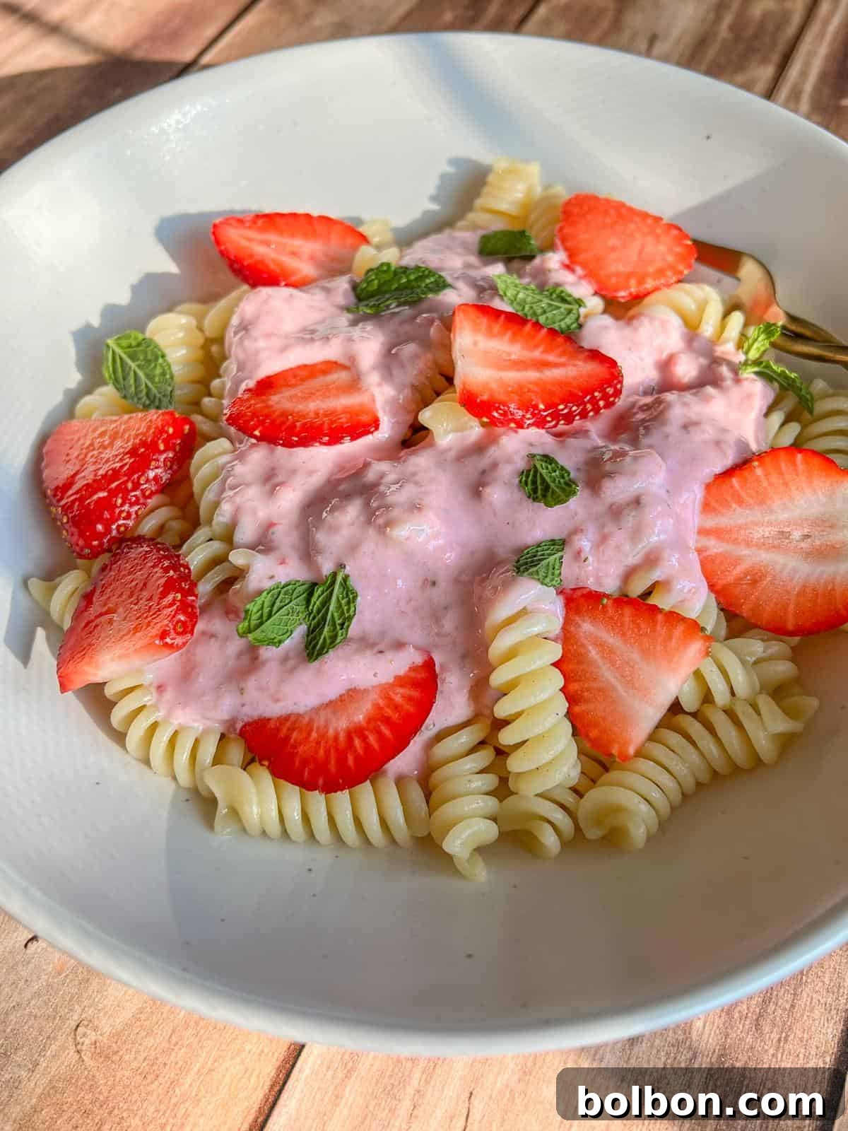 A white plate of fusilli pasta topped with pink strawberry sauce, fresh strawberry slices, and mint leaves, set on a wooden surface in natural light.