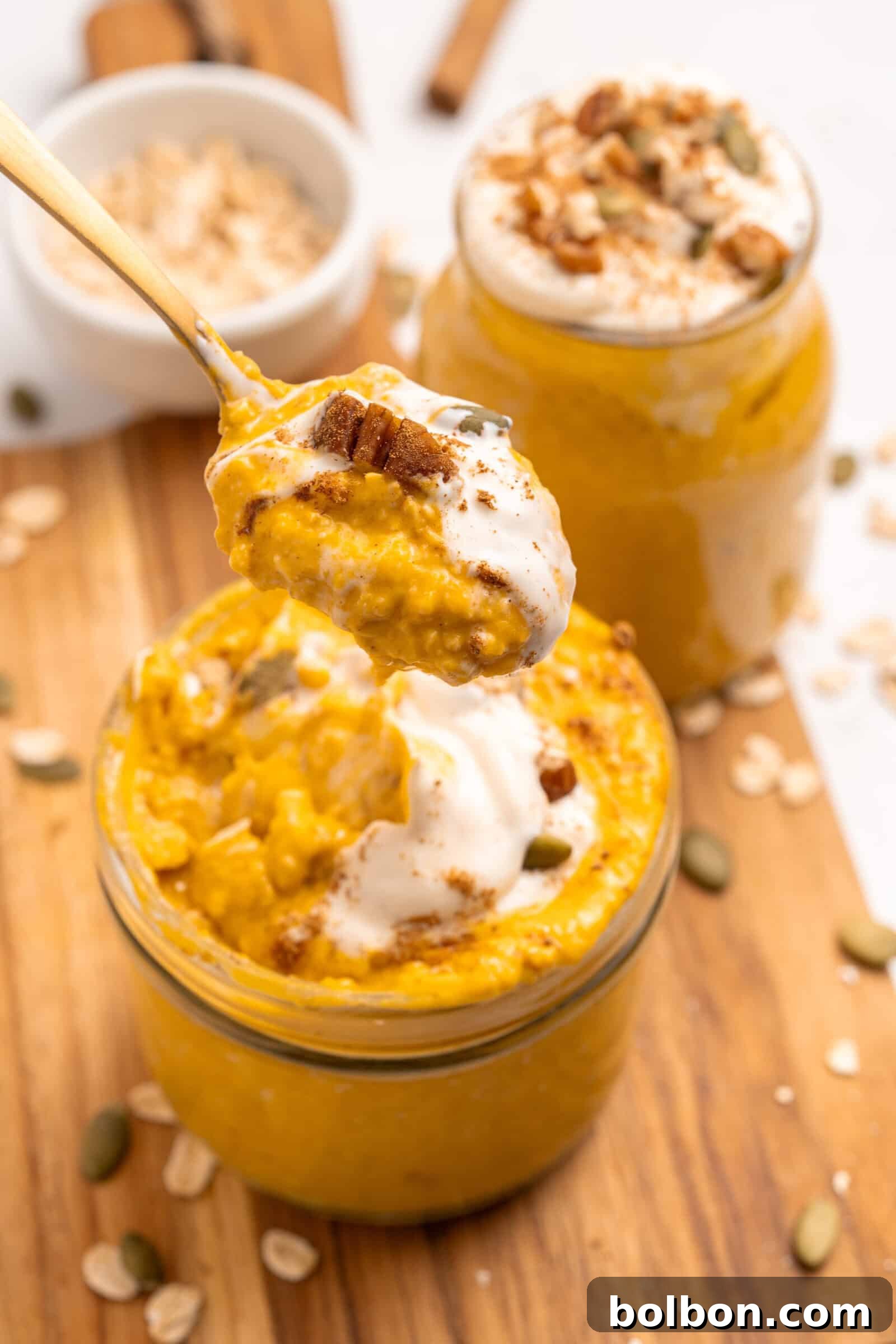 A close-up of pumpkin overnight oats in a jar, topped with cream and nuts, with a spoon lifting a portion. Another jar of oats and scattered oats and seeds are visible on a wooden surface.