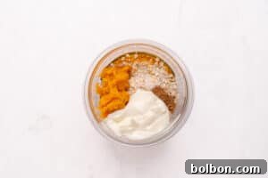 A clear glass bowl containing oats, pumpkin puree, yogurt, and spices, all neatly separated, sits on a white marble surface.