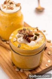 A glass jar filled with creamy orange pumpkin overnight oats, topped with yogurt, pecans, pumpkin seeds, oats, and a sprinkle of cinnamon, sits on a wooden board next to another similar jar.