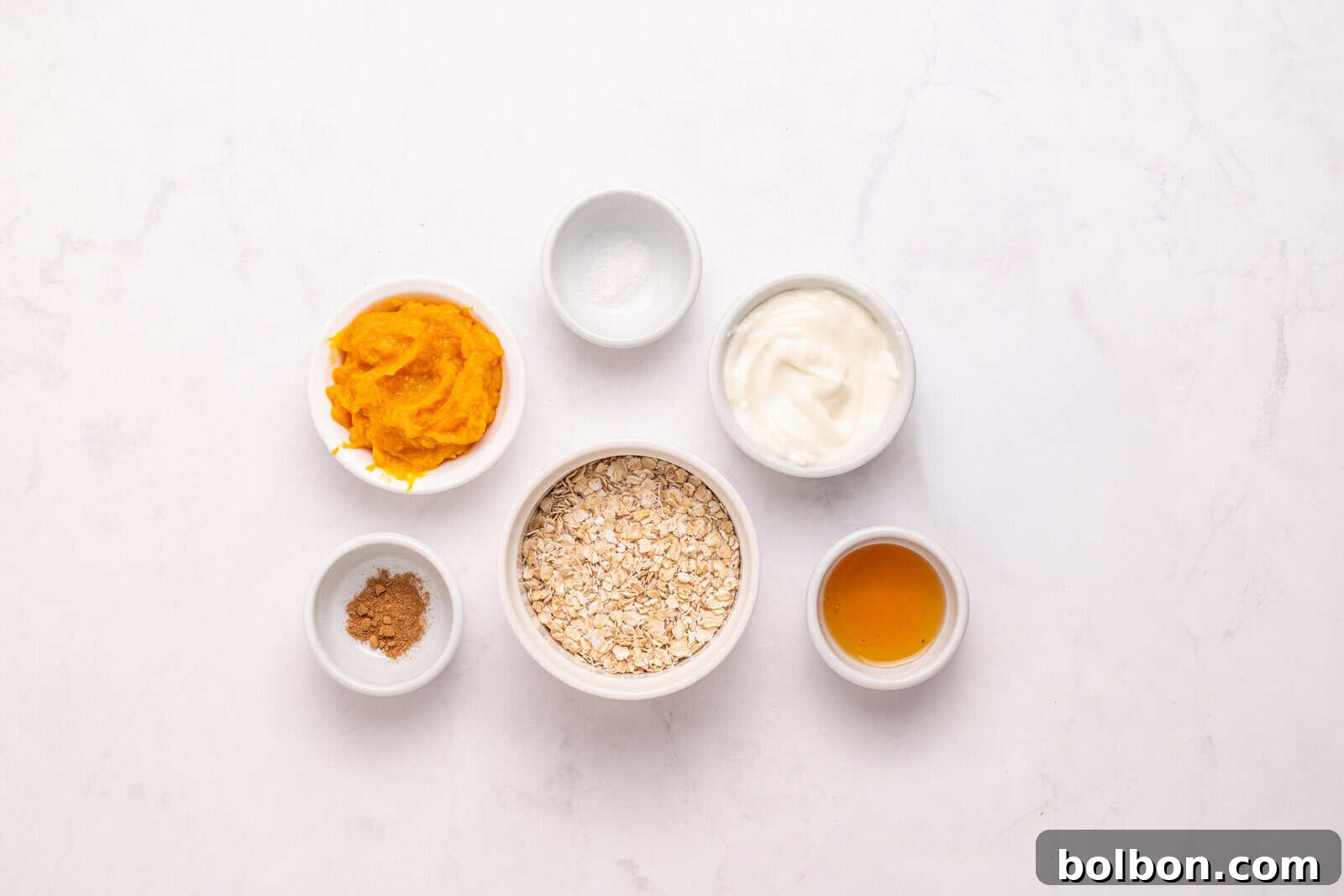 Six small bowls on a white surface, each containing a different ingredient: oats, pumpkin puree, yogurt, maple syrup, ground spice (possibly cinnamon), and a pinch of salt.