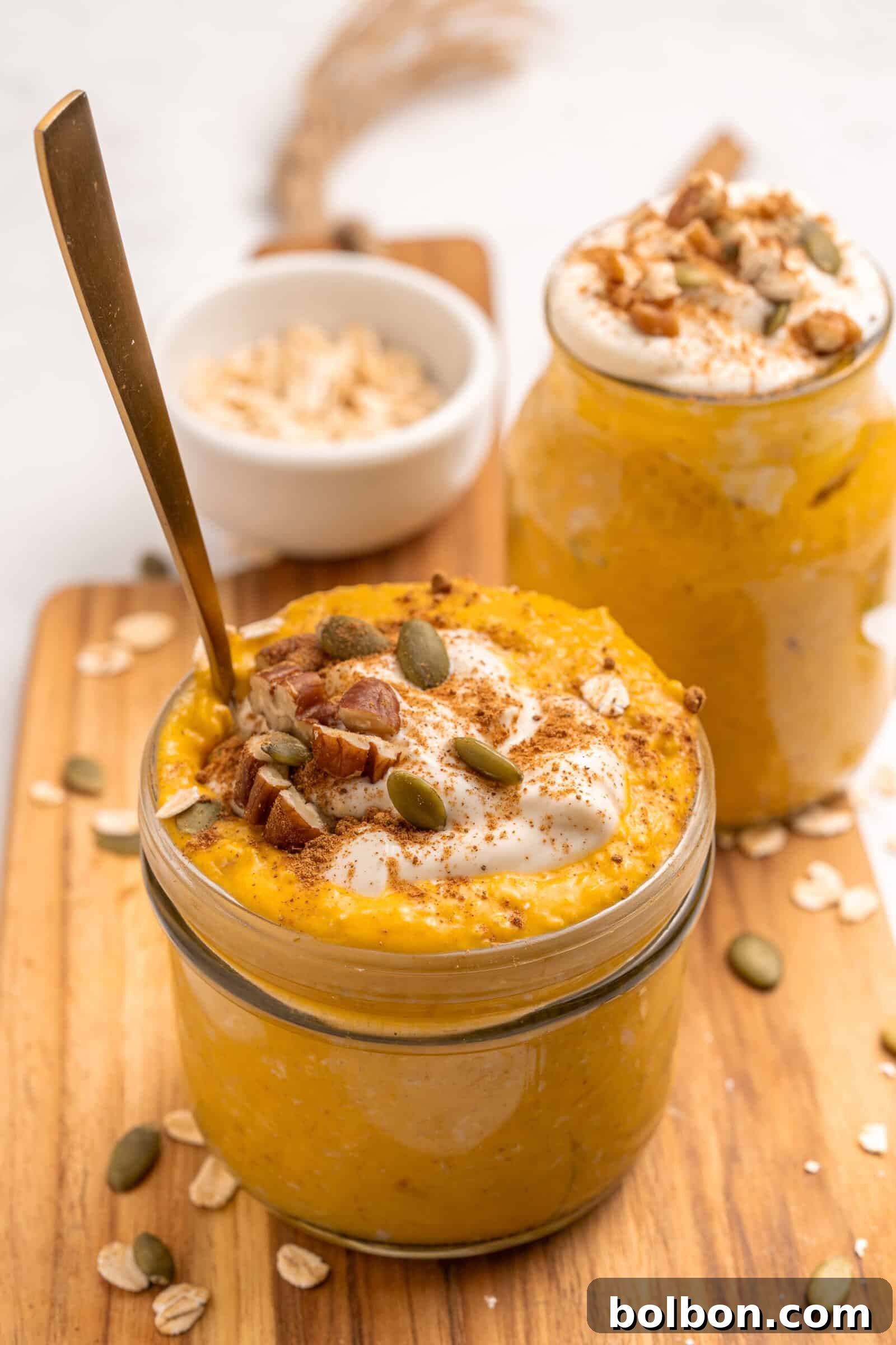 Two glass jars filled with creamy pumpkin overnight oats, topped with yogurt, pecans, pumpkin seeds, and a sprinkle of cinnamon. A spoon is in one jar. Rolled oats are scattered on the wooden board underneath.