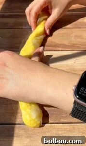 Crispy Rice Paper Burek 7 Hands are twisting a long piece of yellow dough on a wooden surface. One hand wears a smartwatch with a black band. Sunlight casts shadows across the scene.