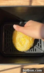 Crispy Rice Paper Burek 9 A hand places a round, sesame-seeded dough into a black air fryer basket, ready for baking. The surface beneath the air fryer is wooden, and sunlight casts soft shadows over the scene.
