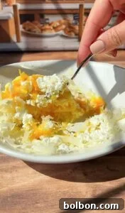 Crispy Rice Paper Burek 4 A hand holding a fork is mixing shredded cheese and crumbled white cheese in a white bowl on a wooden table, with a bakery display blurred in the background.