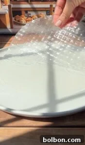 Crispy Rice Paper Burek 5 A hand places a sheet of translucent rice paper onto a white plate, with sunlight casting shadows across the scene and baked goods visible in the background.