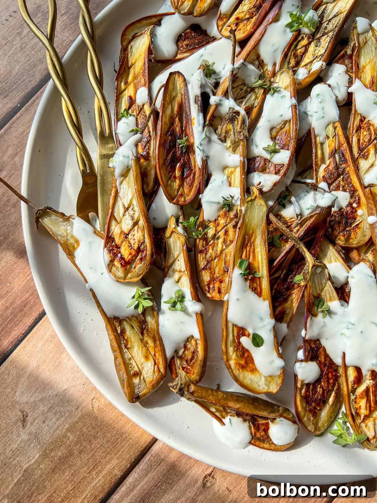 Grilled eggplant halves arranged on a white plate, drizzled with creamy white sauce and garnished with fresh herbs, with gold forks placed on the side.