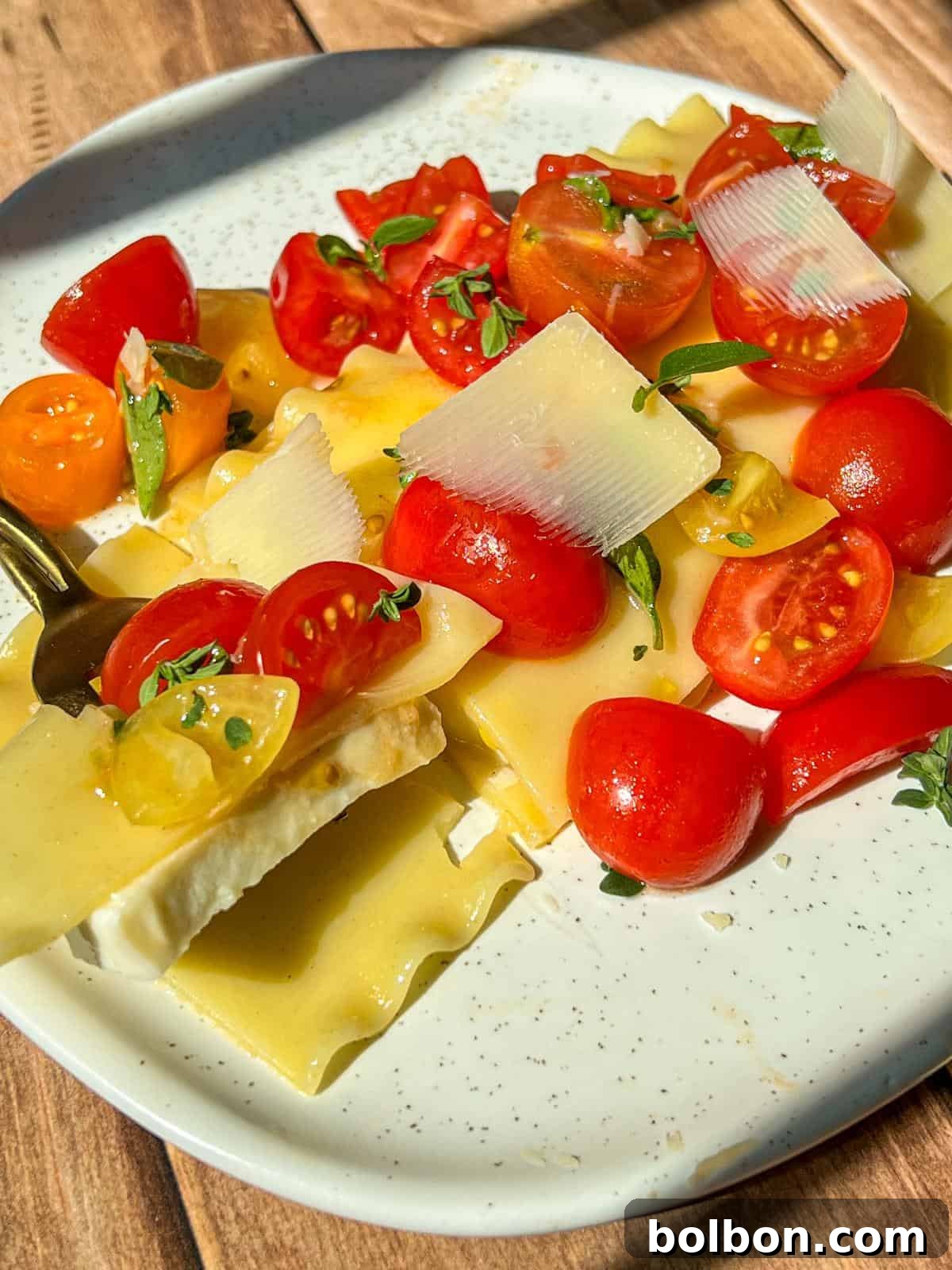 A plate of lasagna sheets topped with halved cherry tomatoes, fresh herbs, and thin cheese slices, all drizzled with olive oil. A fork holds a bite of lasagna, tomatoes, and cheese. Sunlight brightens the dish.