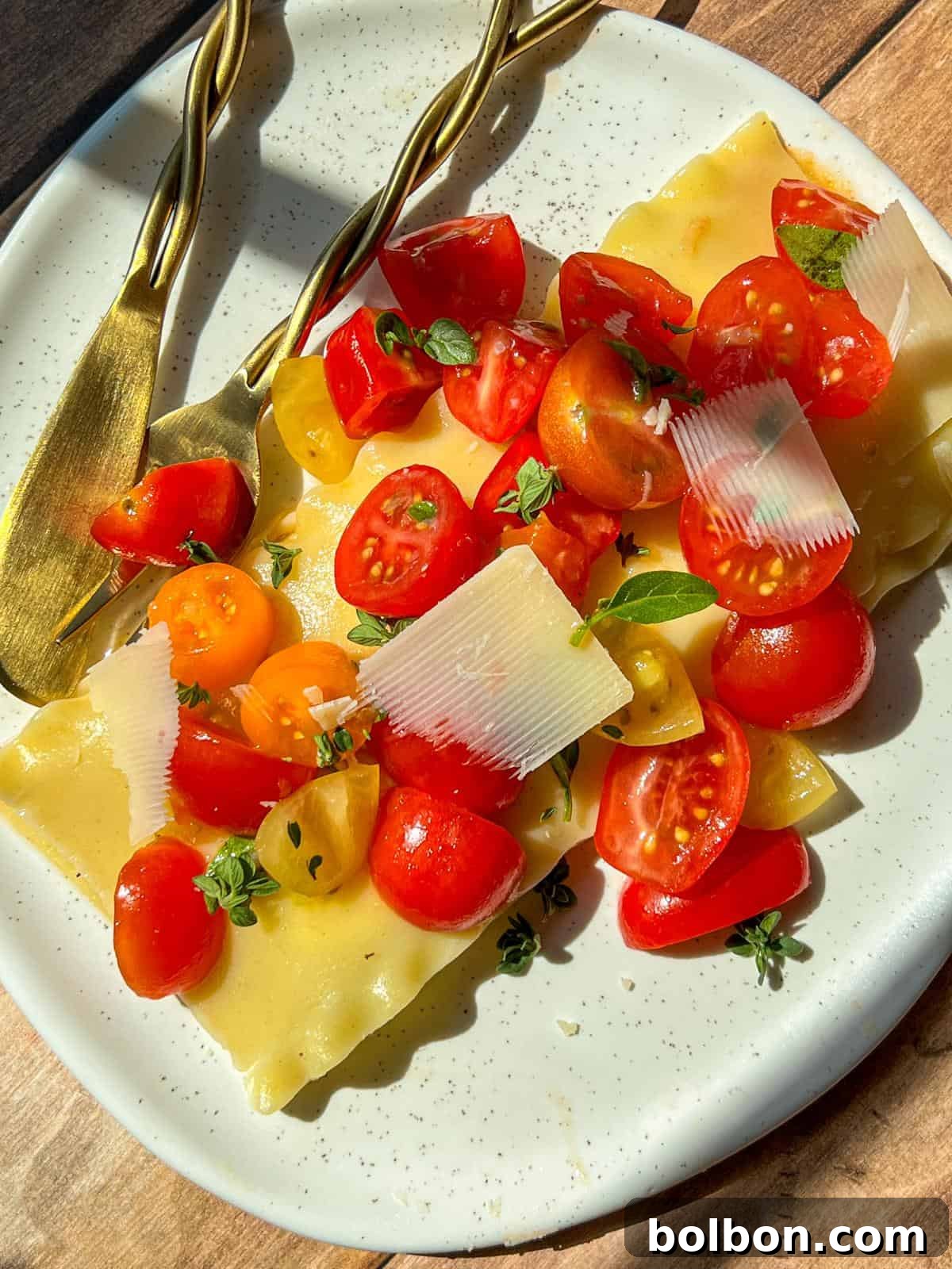 A plate with lasagna sheets topped with cherry tomato halves, fresh herbs, and thin slices of cheese. A gold fork and knife rest on the side, and sunlight casts shadows on the wooden table.