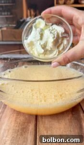 A hand holds a small glass bowl of ricotta cheese above a large glass bowl filled with a frothy yellow mixture on a wooden surface, suggesting a cooking or baking process.