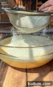 A close-up of flour being sifted through a mesh strainer into a glass bowl filled with a yellow batter, all set on a wooden table with sunlight streaming in.