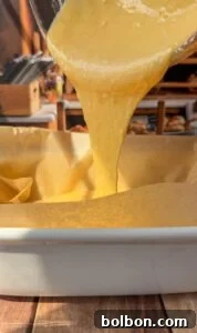 Yellow cake batter is being poured from a bowl into a parchment paper-lined baking dish on a wooden surface, with a warm kitchen setting in the background.