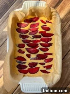 A rectangular baking dish lined with parchment paper holds an unbaked batter topped with neatly arranged slices of red plums, ready to be baked, on a wooden surface.