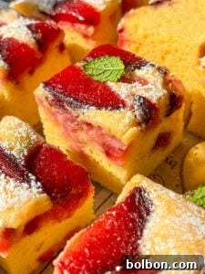 Close-up of several pieces of yellow sponge cake topped with red plum slices and powdered sugar, some garnished with a small green mint leaf. The cake pieces are arranged closely together.