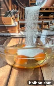 A close-up of sugar being poured from a small glass container into a clear bowl containing three raw eggs on a wooden surface, with a bakery-style kitchen in the background.