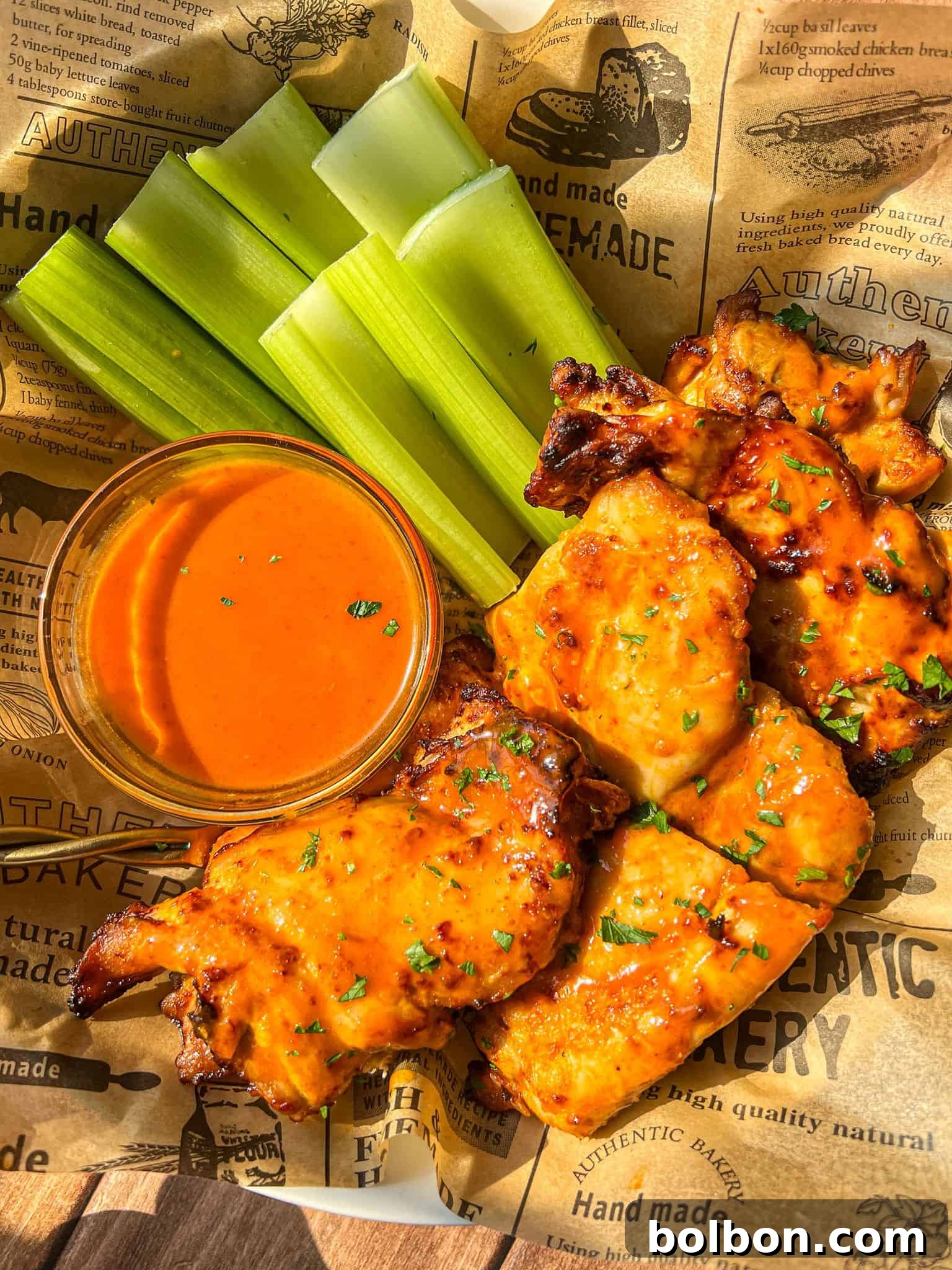 Buffalo chicken wings arranged with celery sticks and a cup of orange buffalo sauce, served on parchment-style paper with text and graphics.