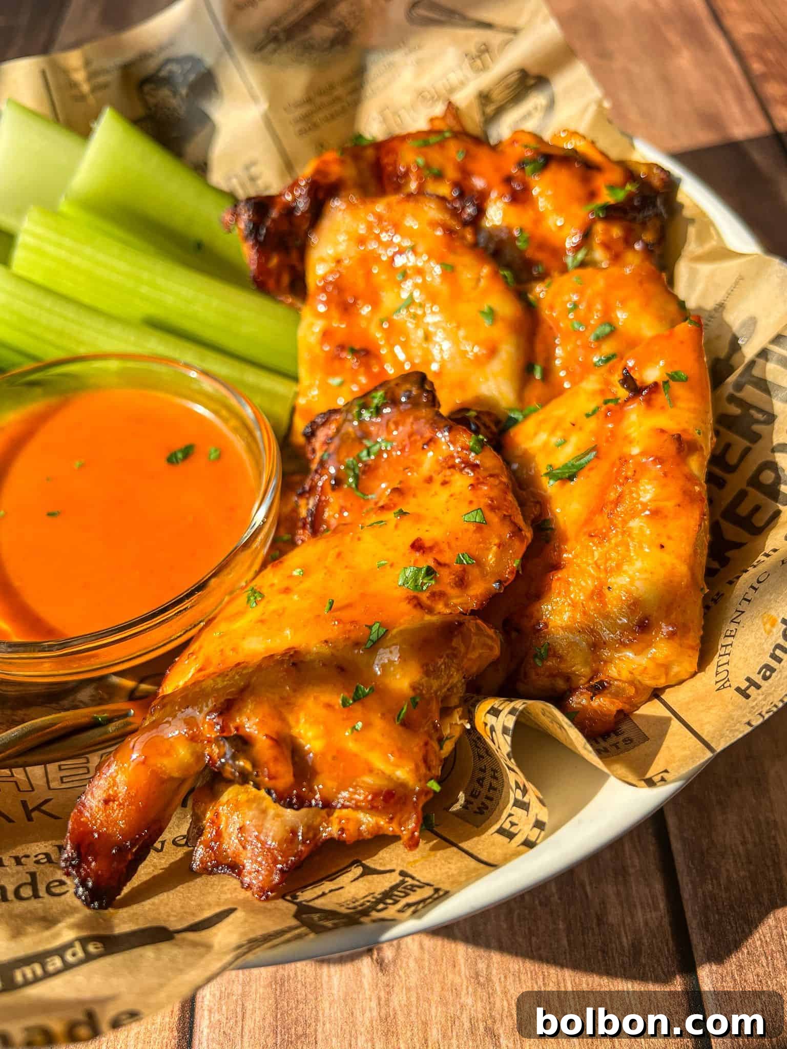 A plate of grilled buffalo chicken wings garnished with herbs, served with fresh celery sticks and a small bowl of orange buffalo sauce on brown parchment paper.